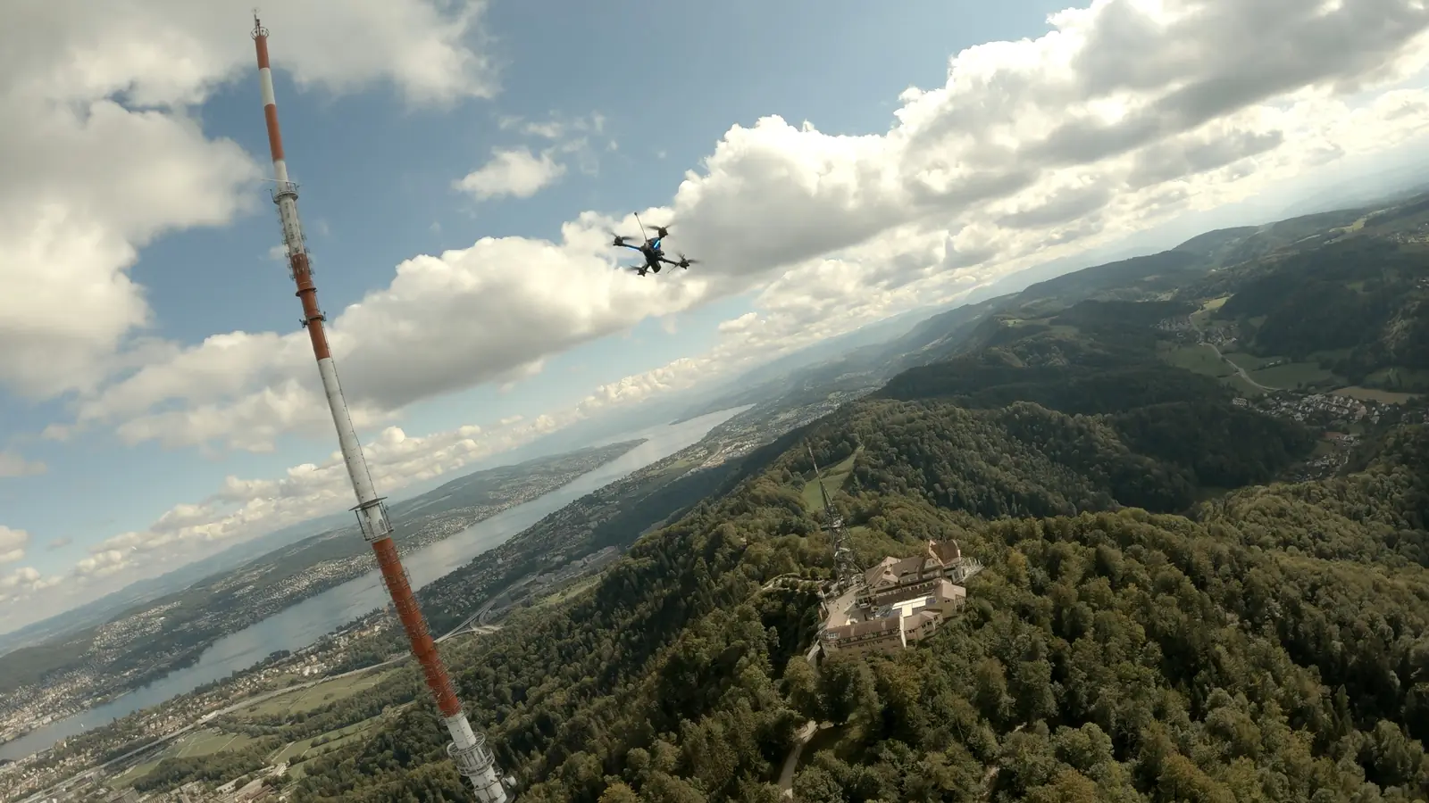 FPV-Aufnahme: eine Drohne fliegt vor einem Sendeturm über einem bewaldeten Hügel.